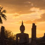 Siluetta di una statua di Buddha e di un chedi tradizionale al tramonto nel Parco Storico di Sukhothai con una palma sullo sfondo